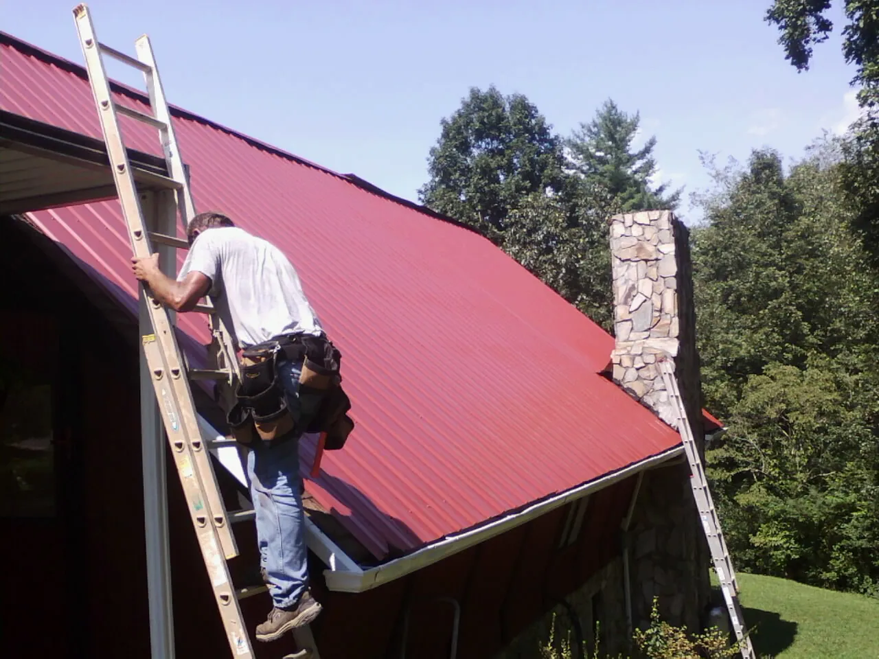 Roofer on a ladder beside a red metal roof and stone chimney — Soffit & Fascia Repair by Roofing Brothers in Lincolnwood